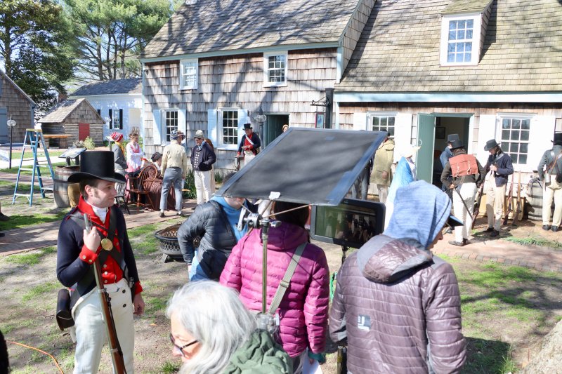 An actor in a soldier’s uniform pauses to watch as director Jenny Alexander, in purple coat, reviews a playback of the “Chaos” scene on a monitor.